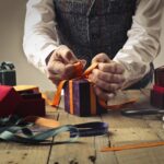 A person wraps a gift with orange ribbon on a rustic wooden table, surrounded by boxes.