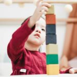 A young child in a red dress plays by stacking colorful wooden blocks in an indoor playroom.