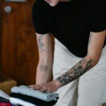 Tattooed woman organizing and packing folded clothes into a suitcase indoors.